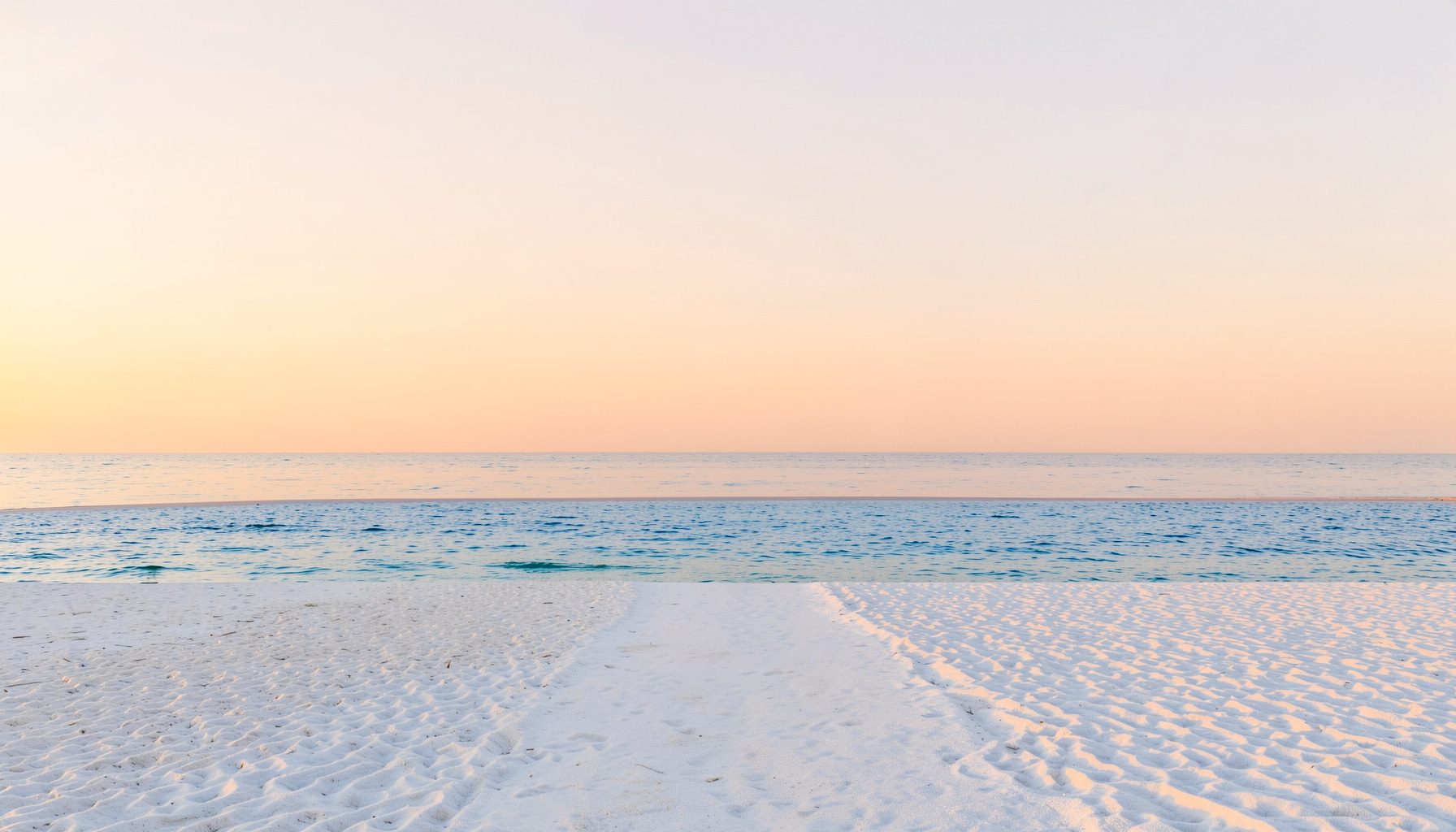 Soft seaside scene with white sand, gentle blue water, and a pale coral sky at golden hour